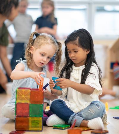 two small girls sitting on a floor building a tower together with magnetic tiles