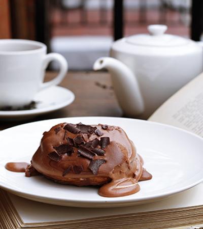 A small chocolate cake sits on a plate on top of an open book. A tea cup and tea pot are in the background.