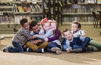 Six children sit on the floor of a library, laughing and hugging