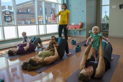 Three mothers lay on yoga mats holding their babies over their heads while an instructor guides them through yoga movements.