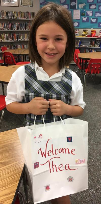 Young girl stands in a library smiling, holding a paper bag that reads "Welcome Thea"
