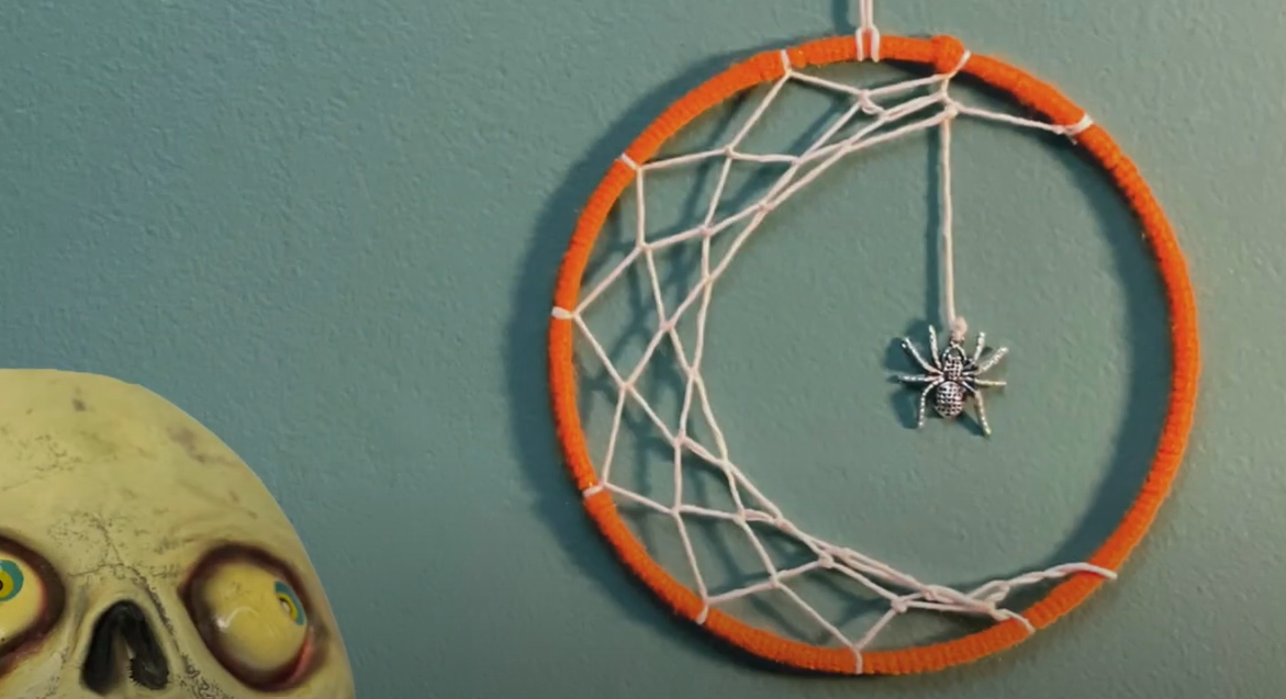 Photograph of a completed spiderweb macrame. The spider is hanging down the center of the orange macrame loop.