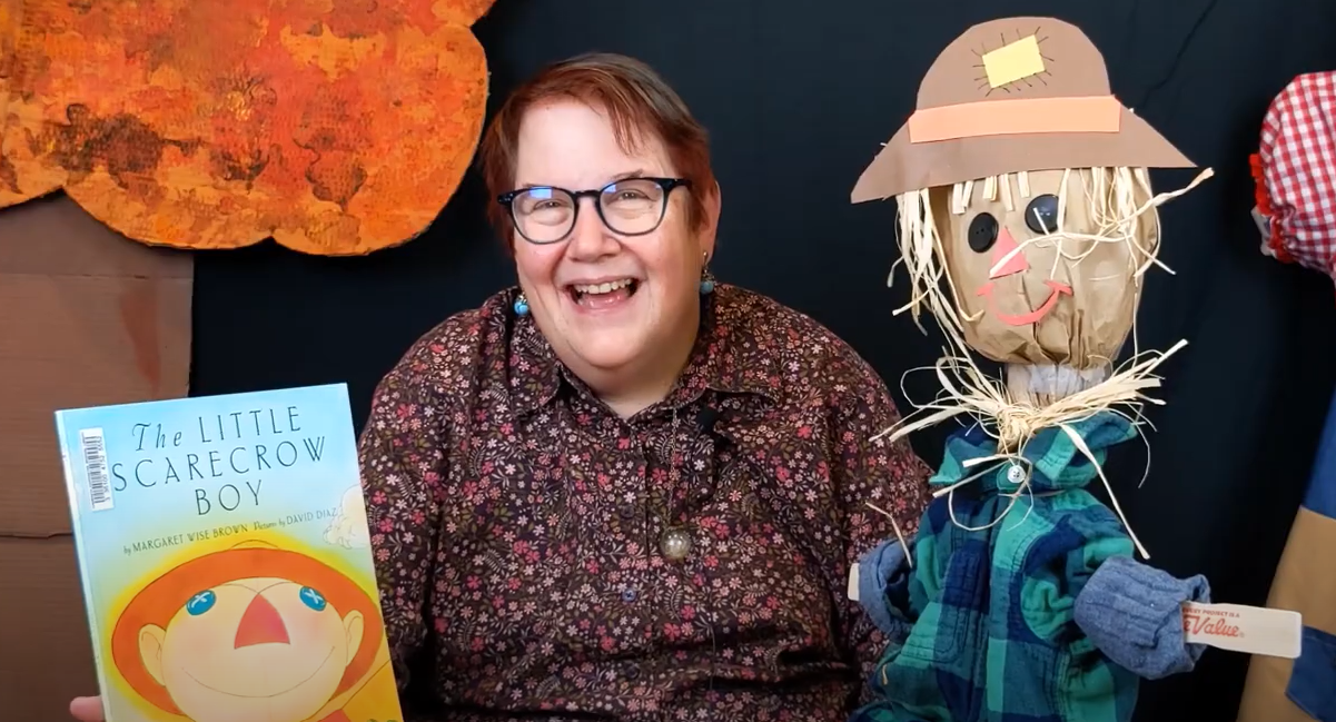 Photograph of librarian smiling next to completed scarecrow book buddy. The librarian is holding the book "The Little Scarecrow Boy" by Margaret Wise Brown