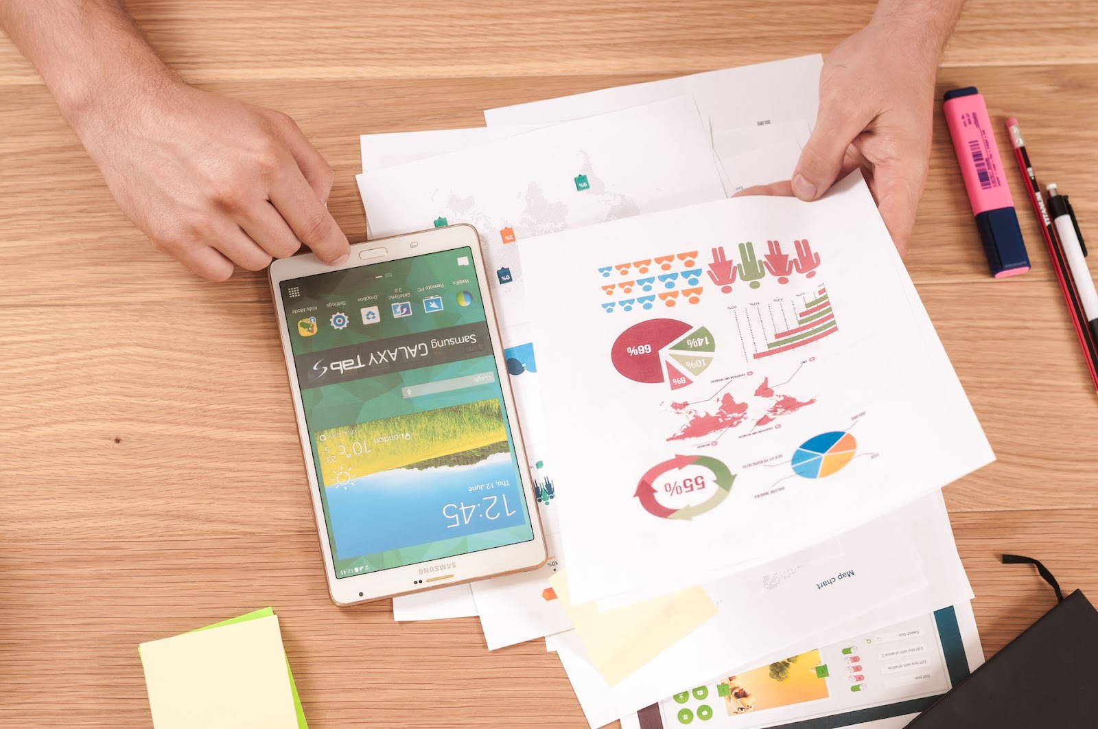 Photograph of a person using a tablet on a table. There is a stack of paper with graphs next to it.