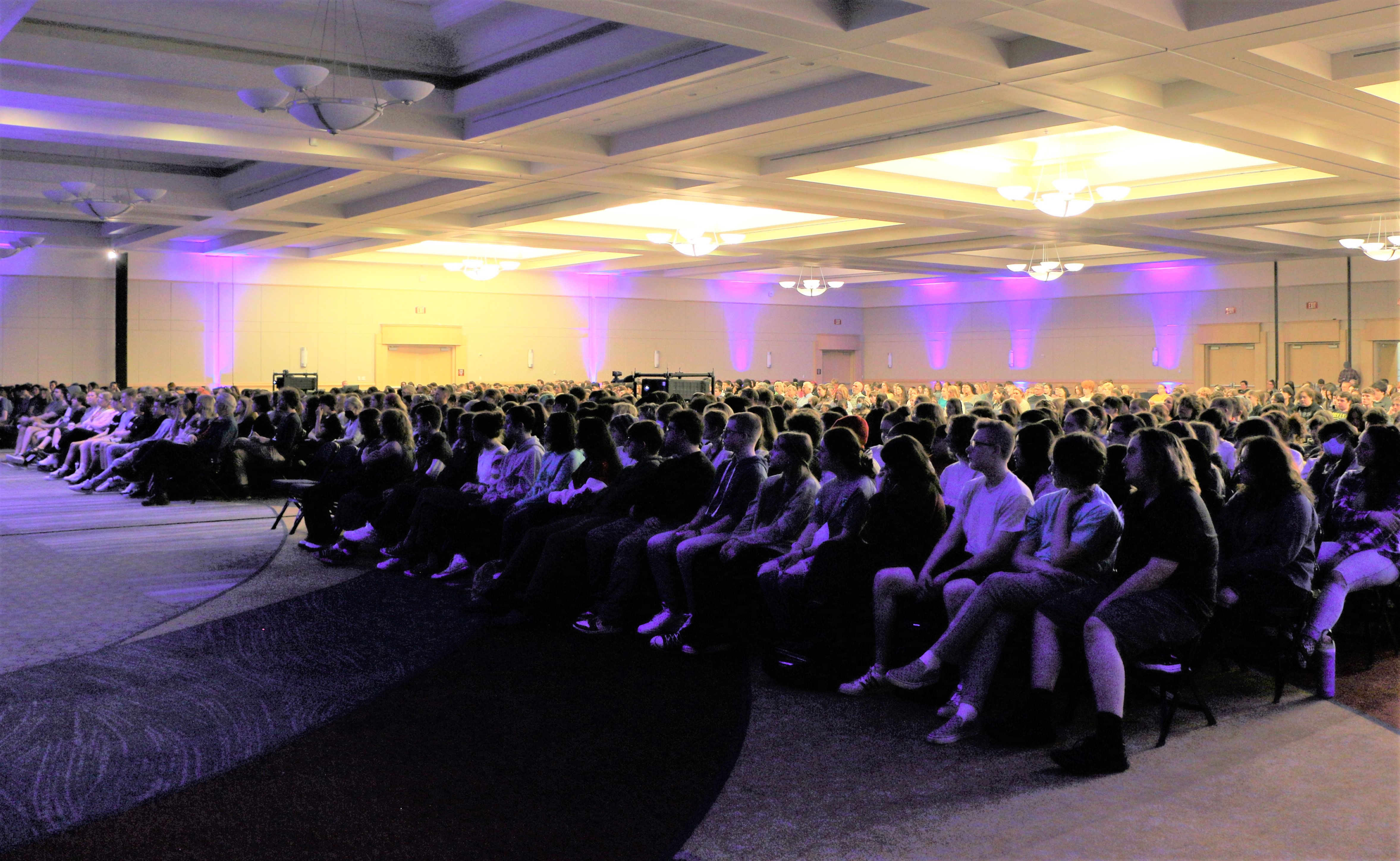 Photograph of a large crowd of people sitting and watching a presentation in a large room.