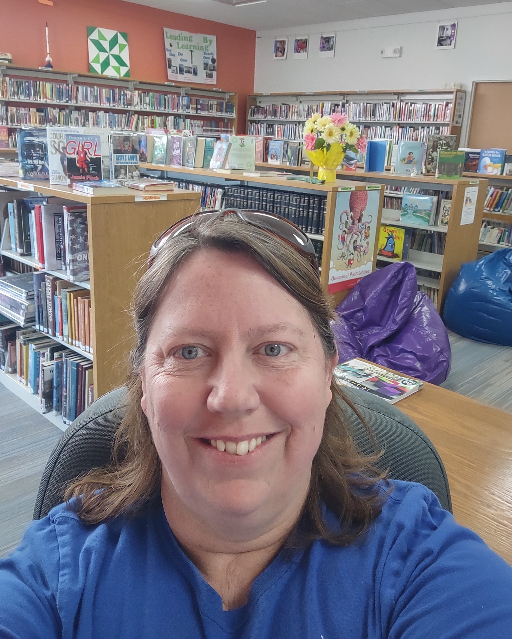 Smiling woman with brown hair and sunglasses on top of her head wearing a blue shirt in a library.