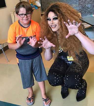 Drag Queen and child face camera and hold hands up in front of them smiling.