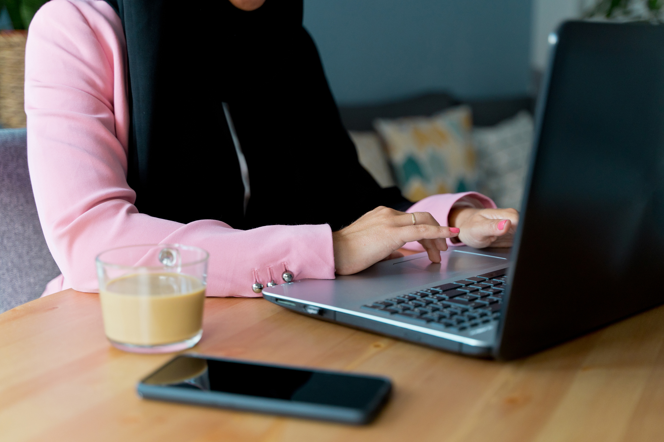 Photograph of person on laptop. The photo is cropped so you cannot see their face. There is a cup of coffee and phone on the table next to the laptop.