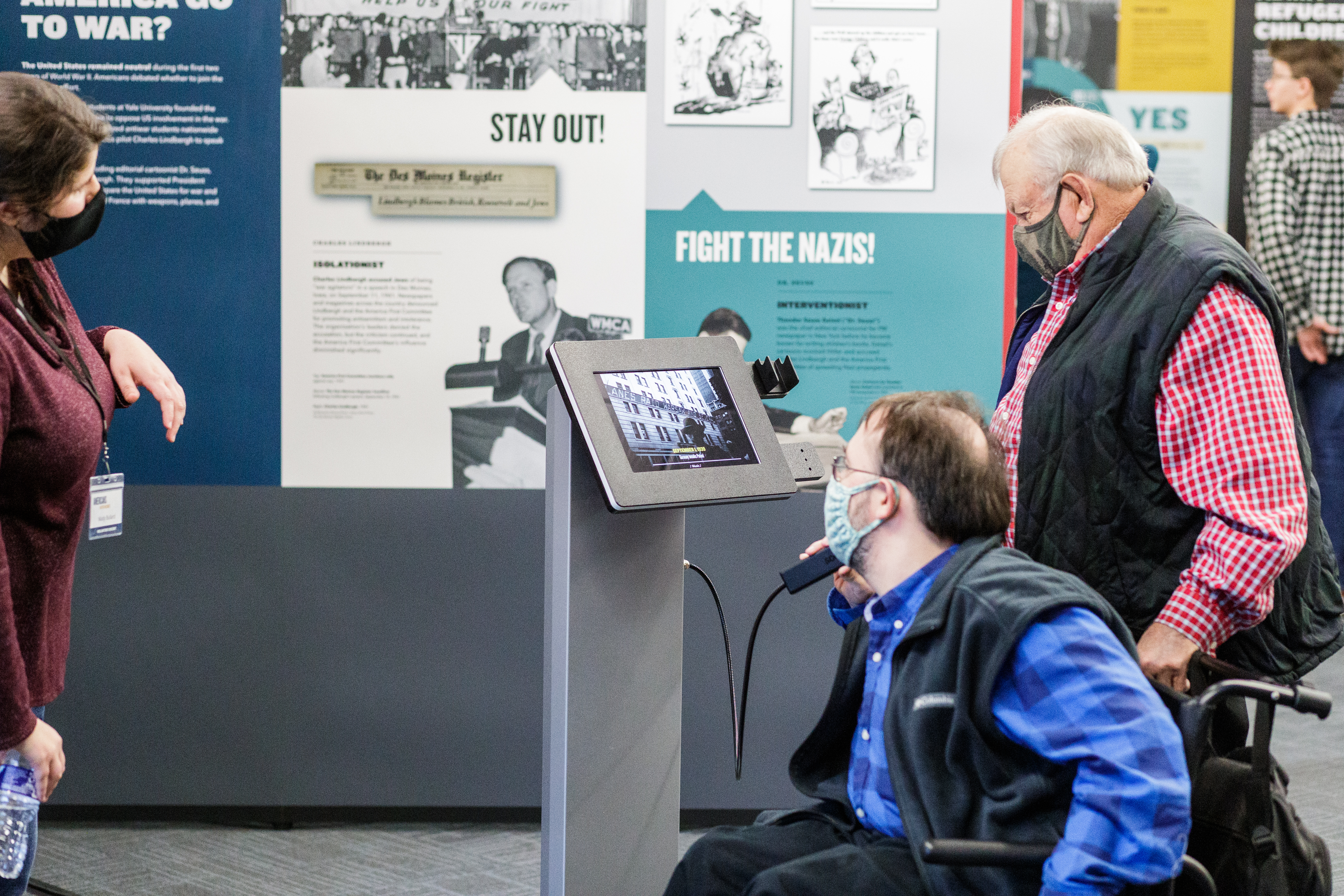 Photograph of a masked person in a wheelchair next to a masked person standing in front of an exhibition panel and interactive iPad for Americans and the Holocaust. There is a masked student docent on the far left of the photo.
