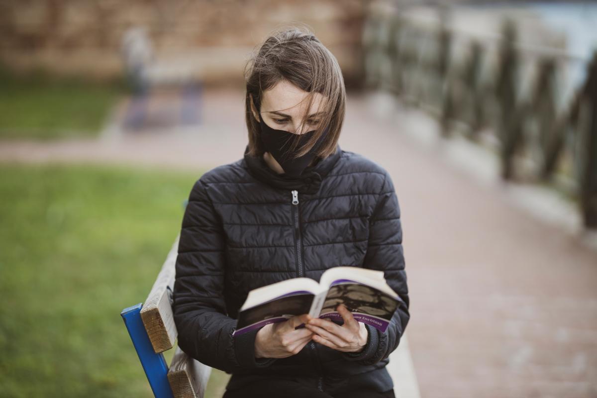 a woman wearing a mask reading on a park bench