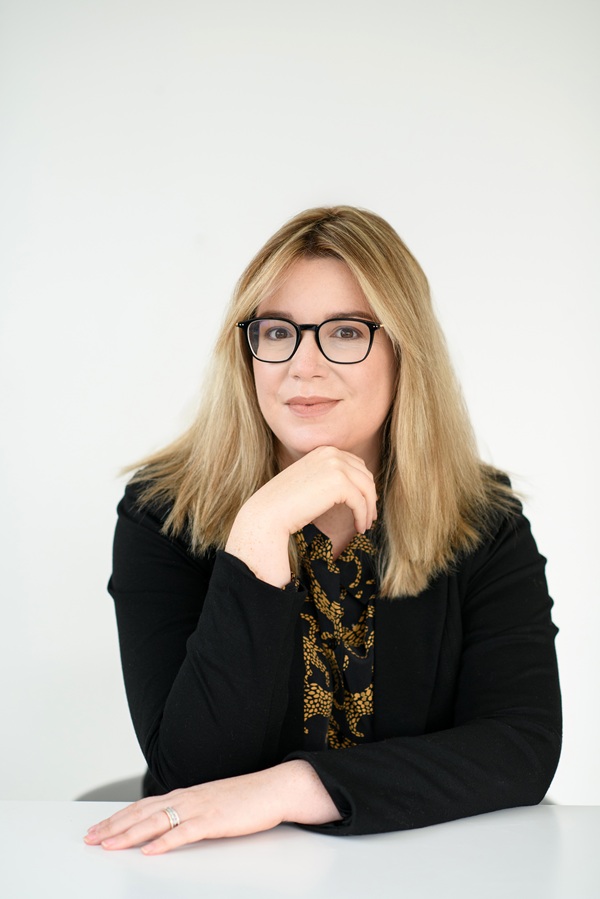 Headshot of Dr. Kate Mercer, a white woman with long blond hair and black rimmed glasses. She is posed with one hand under her chin and another resting on a tabletop.