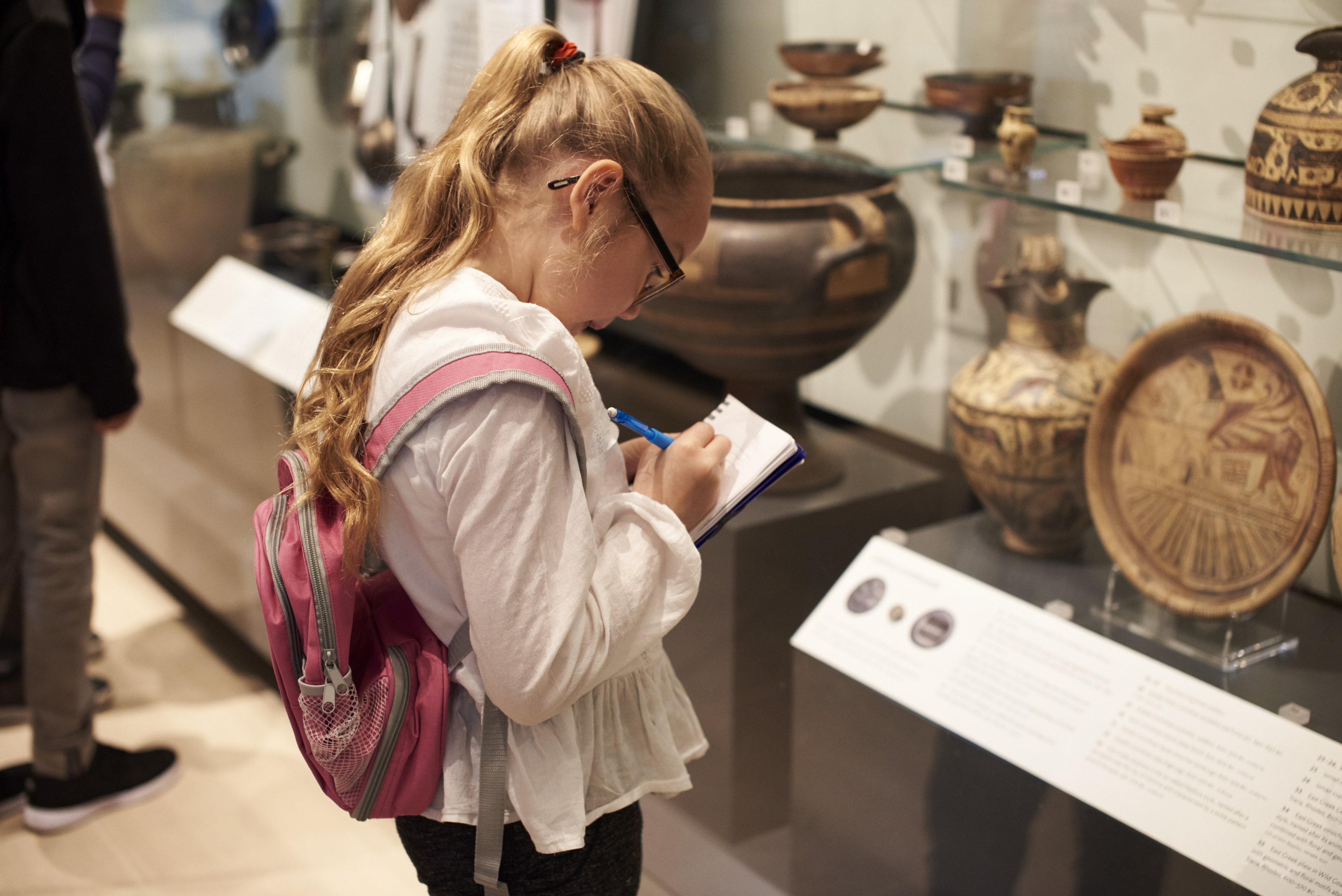 Photo shows a young student taking notes in front of artifacts in a museum case