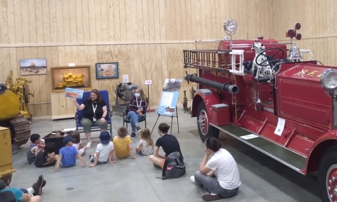 Photograph shows a person reading a book to a group of children sitting on the floor inside a local museum. There is a large, old red fire truck to the right of the group.