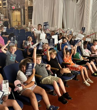 A group of students sitting in rows smile and hold up their passport pamphlets