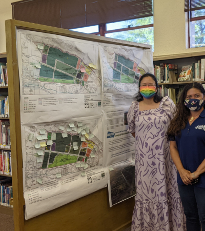Photograph of two masked Waimea librarians standing inside the library in front of a map of the Waimea 400 plan.