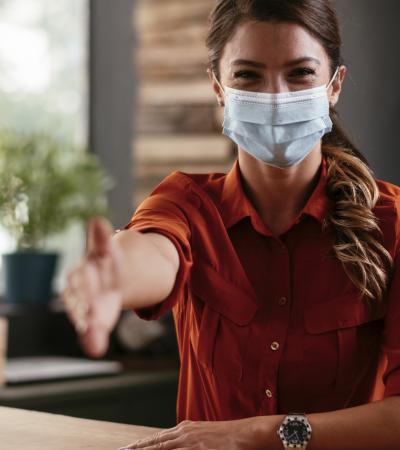 Photograph of woman in medical mask reaching hand out for handshake.
