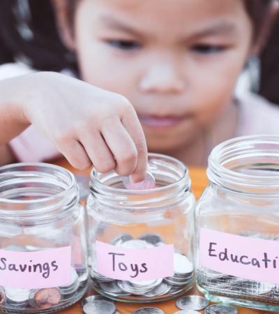 Photograph of child putting coins into three jars. Jars are labeled: Savings, Toys, Education