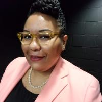 woman with black braided hair in a bun with glasses smiling at camera wearing a pink blazer