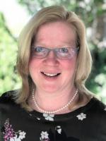 woman with blonde hair and glasses smiling at camera wearing black shirt with white flowers and pearl necklace