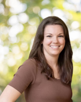 Portrait of a white woman with long brown hair and a light brown t shirt. She is smiling in front of a blurred green foliage background.