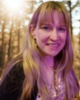 A white woman with long blond hair and bangs is smiling in front of an out of focus forest background. She is wearing a black cardigan and a shimmery top underneath.