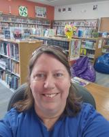 Smiling woman with brown hair and sunglasses on top of her head wearing a blue shirt in a library.