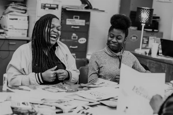Black and white photograph shows two teens laughing at a table with collage materials.