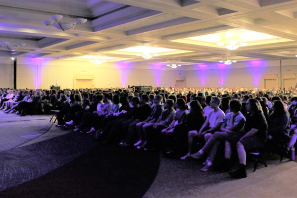 Photograph of a large crowd of people sitting and watching a presentation in a large room.