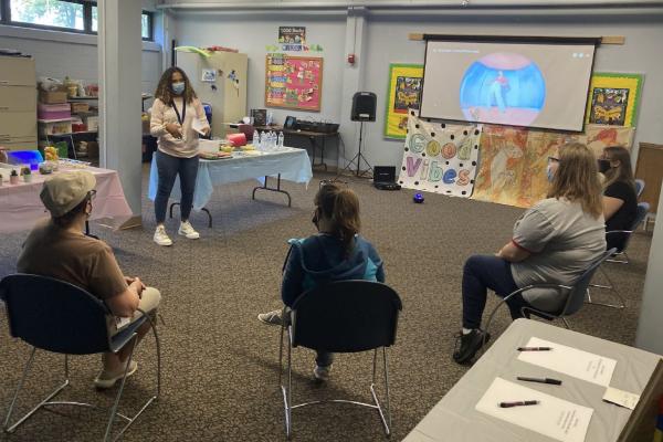 Photograph of intern Hope Campbell standing in front of five library staff members. Hope is presented on mental health. All people are masked and there is a table of snacks and a presentation playing on the projector.