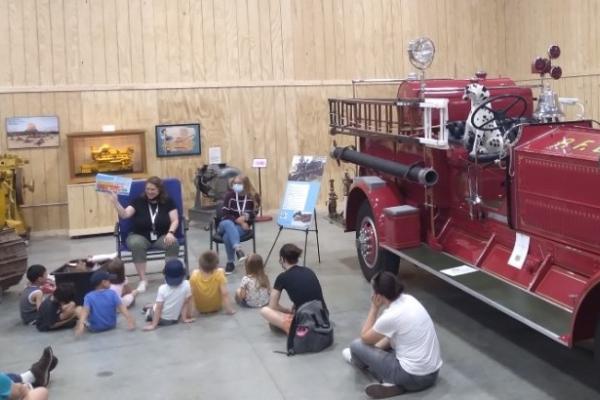 Photograph shows a person reading a book to a group of children sitting on the floor inside a local museum. There is a large, old red fire truck to the right of the group.