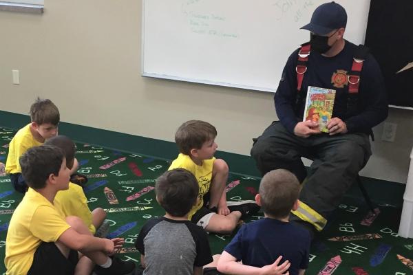 Photograph shows a firefighter reading a book to a group of children.