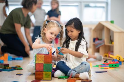two small girls sitting on a floor building a tower together with magnetic tiles