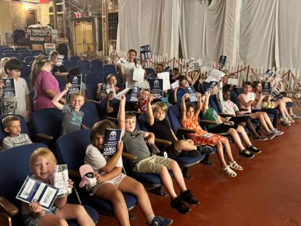 A group of students sitting in rows smile and hold up their passport pamphlets