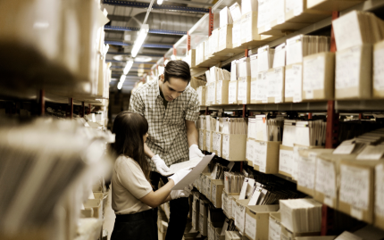 Photograph of two people looking through boxes of archived materials on bookshelves.