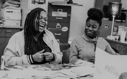 Black and white photograph shows two teens laughing at a table with collage materials.