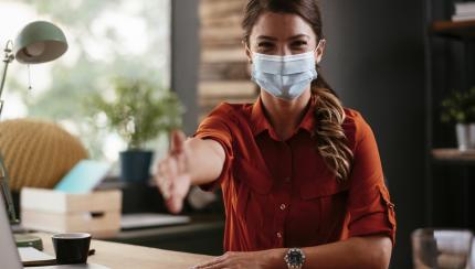 Photograph of woman in medical mask reaching hand out for handshake.