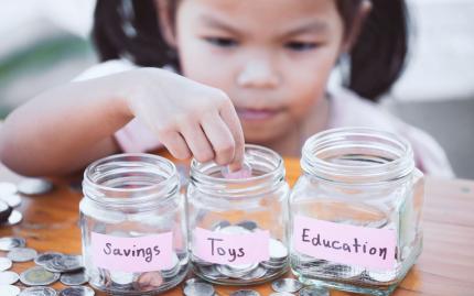 Photograph of child putting coins into three jars. Jars are labeled: Savings, Toys, Education