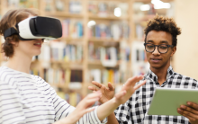 Photograph of two people in a library. The person on the left is wearing a virtual reality headset, the person to their right does not have a headset on and is reading out loud from a book to guide the other person.