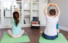 Photograph of two kids sitting on green yoga mats facing an open laptop screen. They have their arms above their heads in a sitting yoga pose.