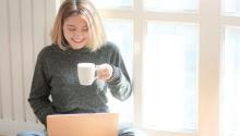 A photo of a woman smiling down at her laptop with coffee in her hand.