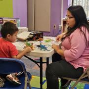 A child and caretaker play with toy dinosaurs