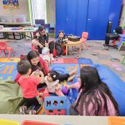 A group of toddlers and caretakers play on brightly colored floor mats