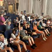 A group of students sitting in rows smile and hold up their passport pamphlets