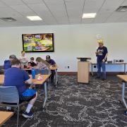 Pokemon Day room set up with people sitting at tables. A person is standing at a podium with a Pikachu hat.