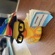 A rubber duck with glasses and a rainbow bow holding a small book with the text Stress ReDUCKtion