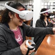 Photograph of two students sitting at a table in the library wearing VR headsets and interacting with the game.
