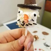Hand holding a marshmallow on a stick, with decorations to make it look like a snowman.