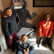 Three kids stand and smile in front of superhero cardboard cutout