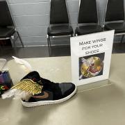Photograph of a craft station - "Make wings for your shoes." There is a black sneaker with gold wings attached to it set out on the table.