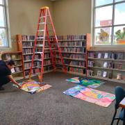 Photograph of final panel art pieces being installed in the library Teen Room. There are bookshelves, windows, a ladder, and the panels ready to be installed on the floor.